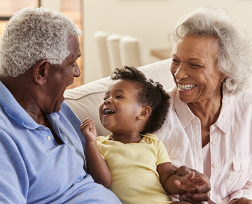 Grandparents laughing with a baby on a sofa.